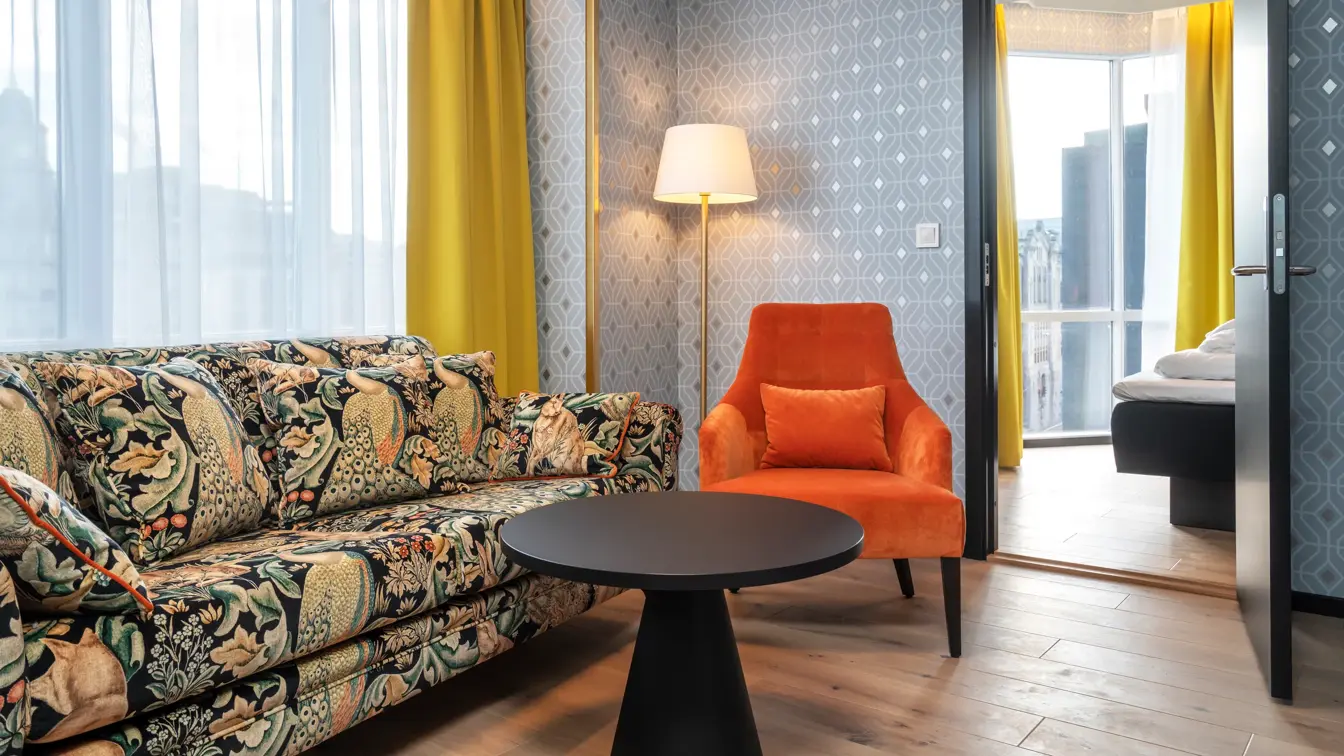 Mønstret fargerik sofa, rundt sort bord, oransje stol, mønstret sølv tapet og gule gardiner i stue på junior suite på Thon Hotel Cecil