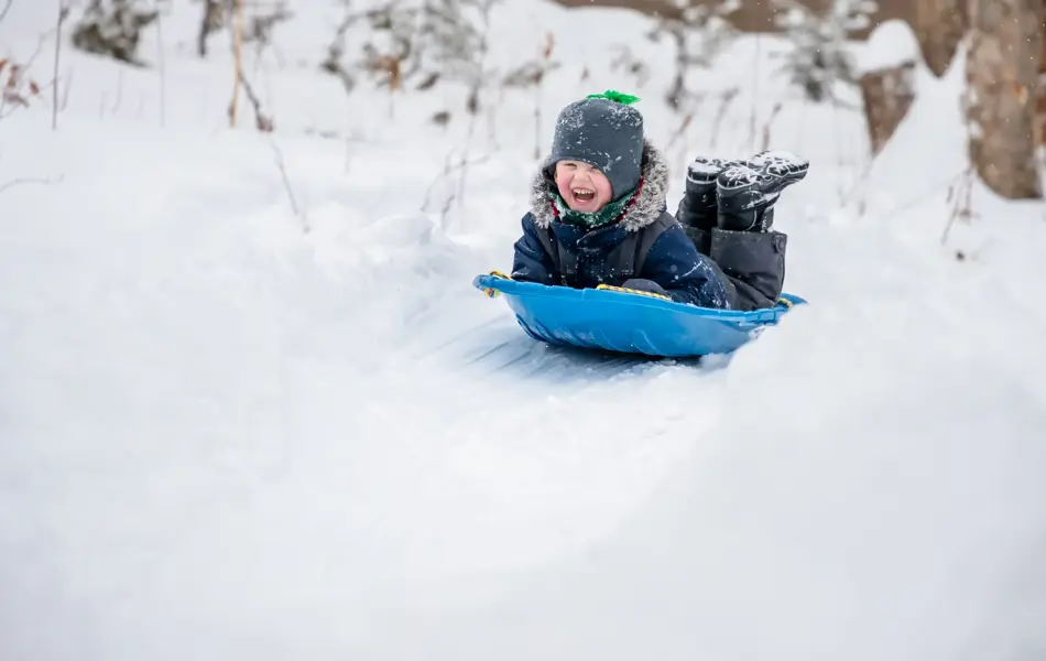 Little Boy Sliding in the Snow Outdoors in Winter. He is smiling and having a lot of fun.