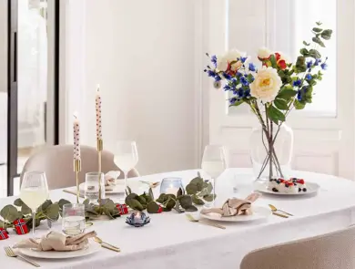 Bord dekket til 17. mai, hvit duk, vase med blomster, grønne grener på bordet, lysestaker, hvitt service, linservietter og vinglass.