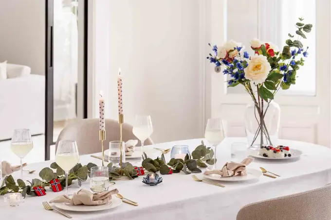 Bord dekket til 17. mai, hvit duk, vase med blomster, grønne grener på bordet, lysestaker, hvitt service, linservietter og vinglass.