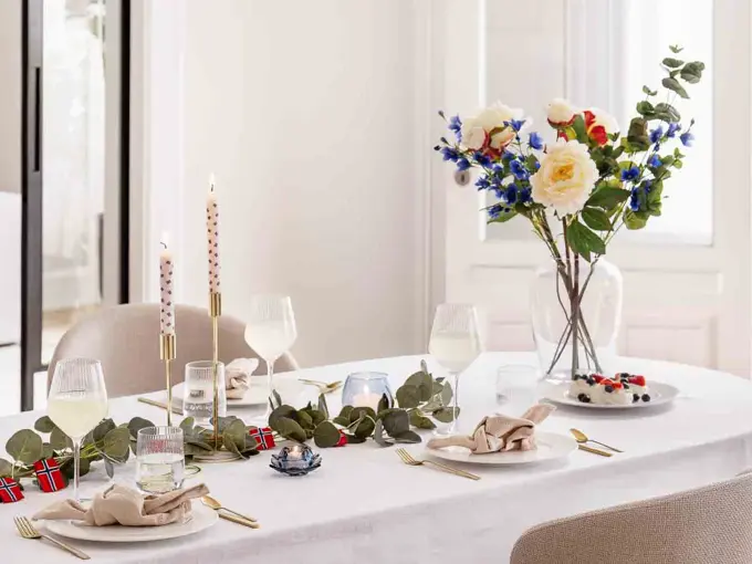 Bord dekket til 17. mai, hvit duk, vase med blomster, grønne grener på bordet, lysestaker, hvitt service, linservietter og vinglass.