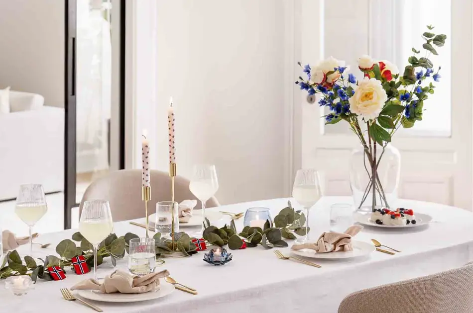 Bord dekket til 17. mai, hvit duk, vase med blomster, grønne grener på bordet, lysestaker, hvitt service, linservietter og vinglass.