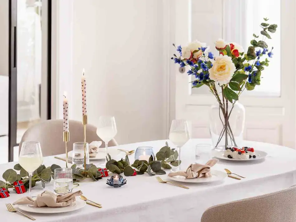 Bord dekket til 17. mai, hvit duk, vase med blomster, grønne grener på bordet, lysestaker, hvitt service, linservietter og vinglass.