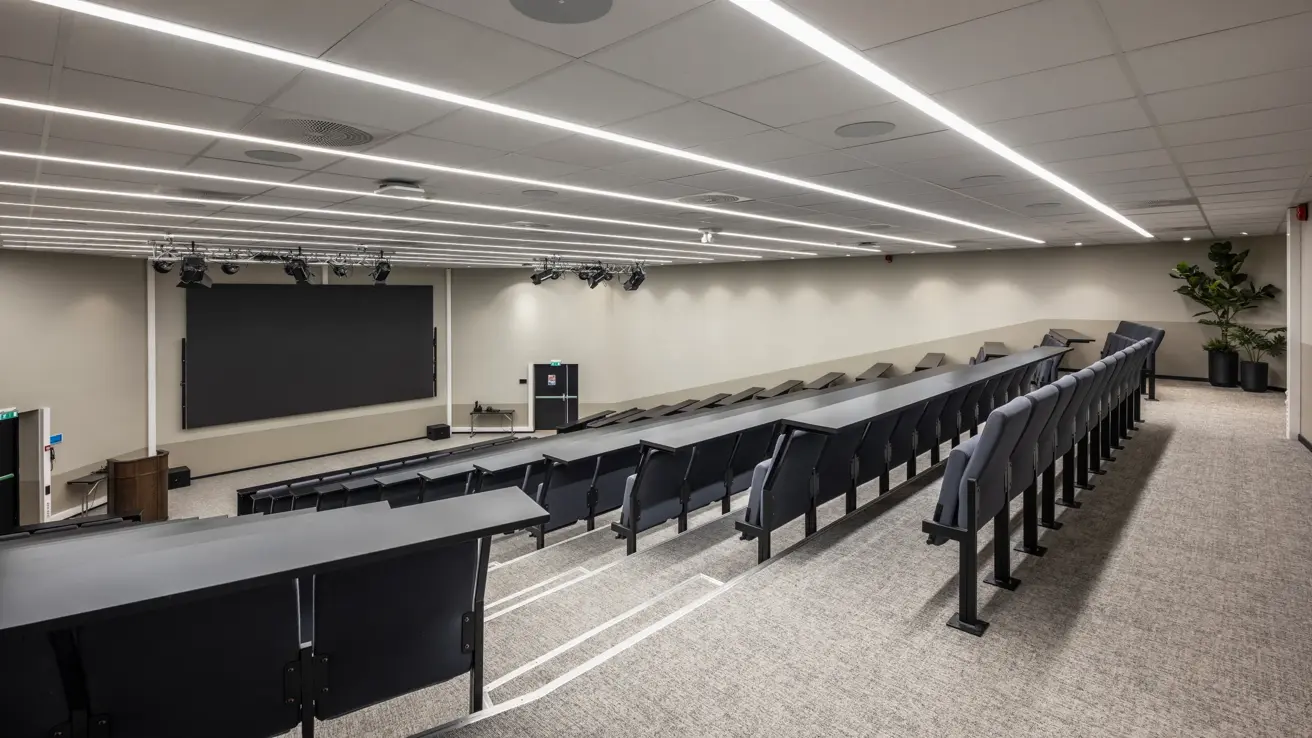 Stort auditorium sett fra den ene siden bak i lokalet med stor skjerm foran, to utganger og talerstol.