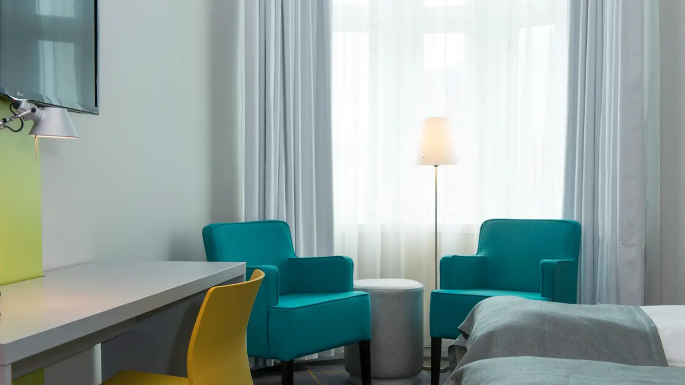 Nærbilde av Standard Twin Room på Thon Hotel Trondheim. Grått skrivebord, gul kontorstol, to turkise lenestoler, to enkeltsenger, vegghengt TV. Store vinduer gir mye godt lys.