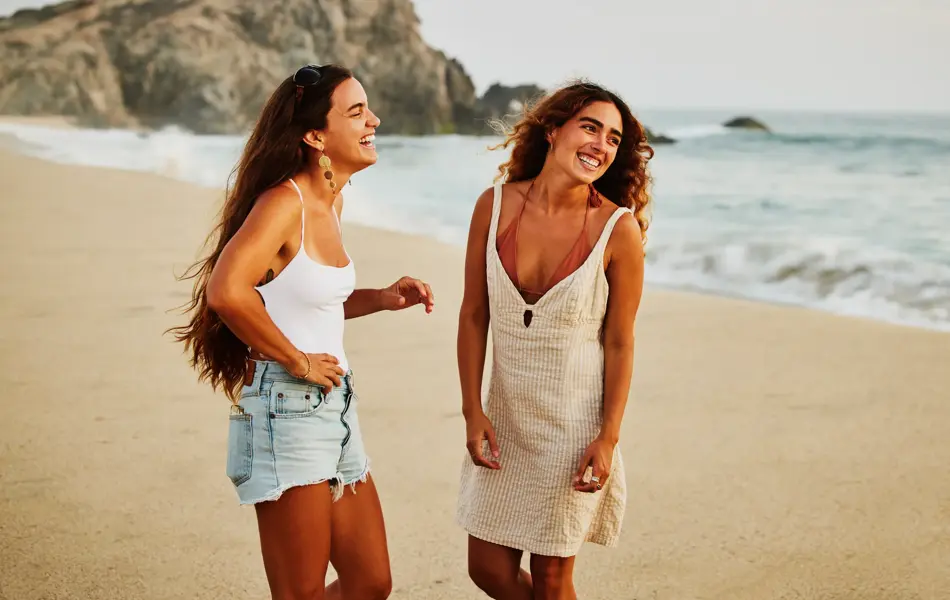 Medium wide shot of laughing female friends hanging out on tropical beach at sunset while on vacation