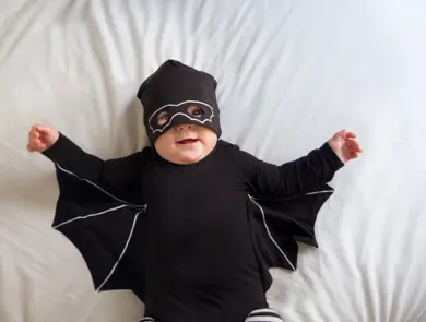 Baby boy is laying in bed wearing a mask and a bat costume while looking at the camera and smiling
