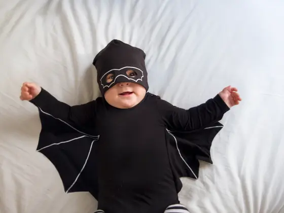 Baby boy is laying in bed wearing a mask and a bat costume while looking at the camera and smiling