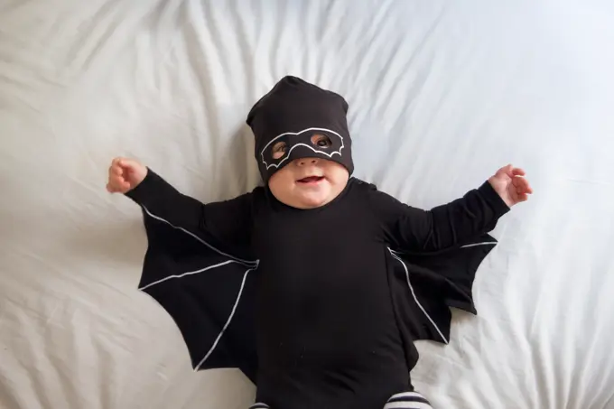 Baby boy is laying in bed wearing a mask and a bat costume while looking at the camera and smiling