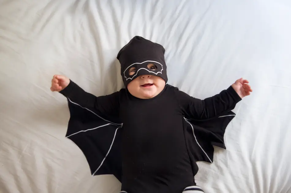 Baby boy is laying in bed wearing a mask and a bat costume while looking at the camera and smiling