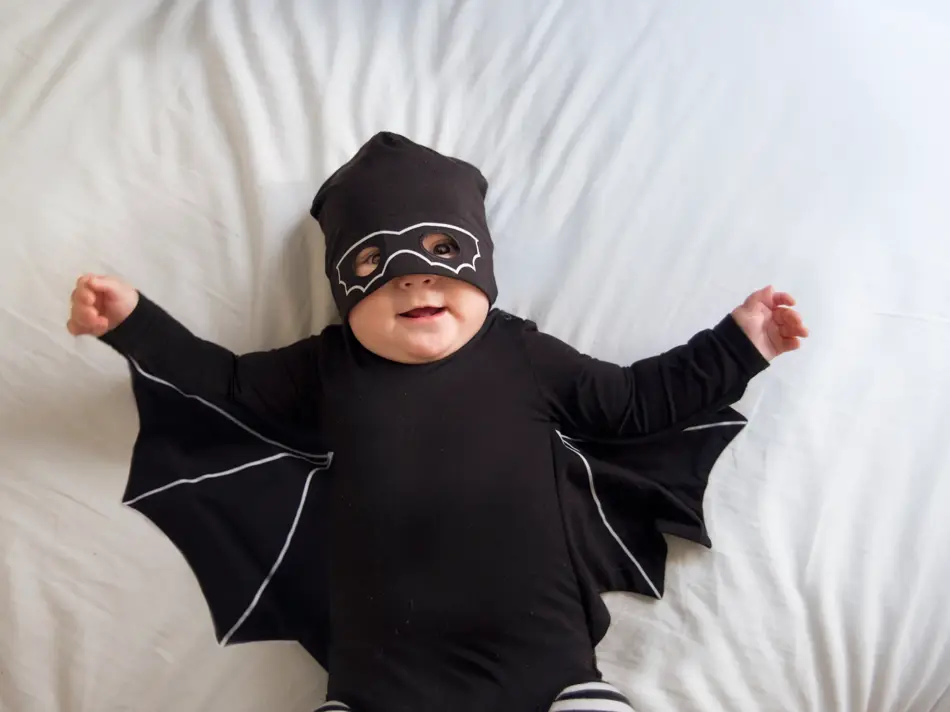 Baby boy is laying in bed wearing a mask and a bat costume while looking at the camera and smiling
