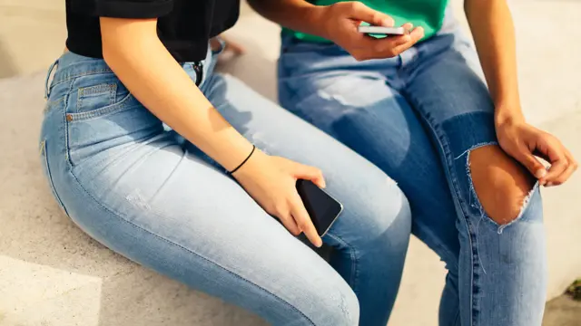 Utsnitt av to jenter i jeans som sitter med mobilen