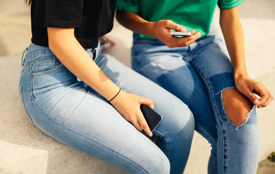 Utsnitt av to jenter i jeans som sitter med mobilen