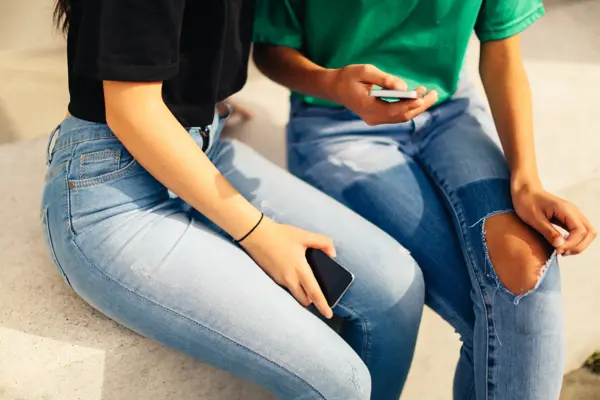 Utsnitt av to jenter i jeans som sitter med mobilen