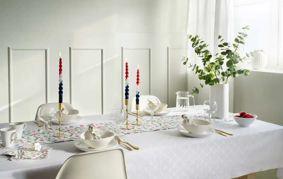 Langbord dekket med hvit duk, løper med blomster, vase med blomster, hvitt service og lysestaker.