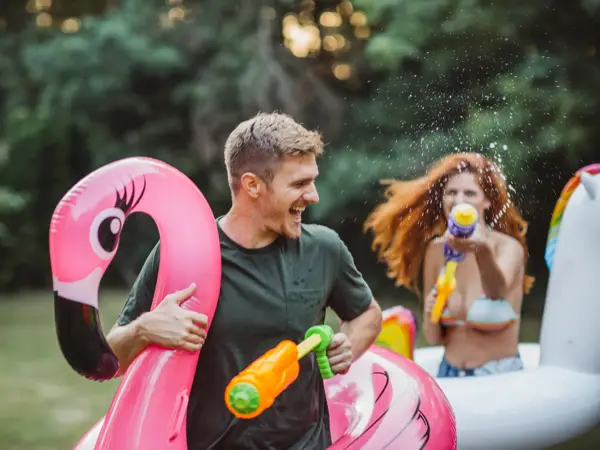 Young couple having fun with water guns, floaty unicorn and flamingo in thair backyard. Enyouing in hot summer days.