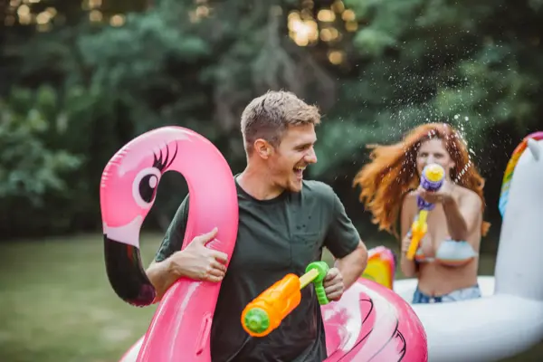 Young couple having fun with water guns, floaty unicorn and flamingo in thair backyard. Enyouing in hot summer days.