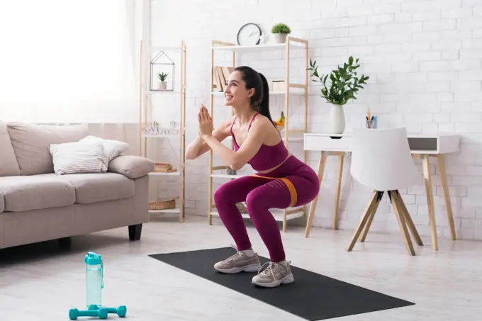 Stay home strength training. Beautiful girl doing squats with rubber band in light room. Copy space