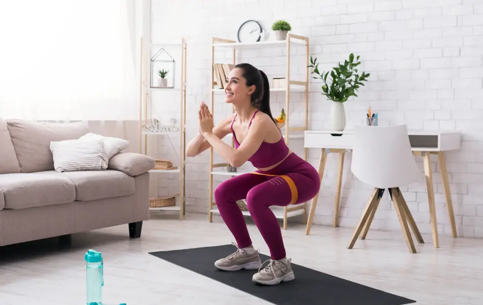 Stay home strength training. Beautiful girl doing squats with rubber band in light room. Copy space