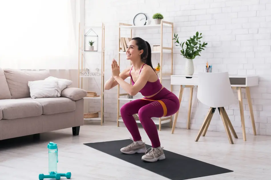 Stay home strength training. Beautiful girl doing squats with rubber band in light room. Copy space