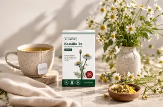 En boks med kammille te fra Kloster står ved siden av en kopp med te og en vase med blomster.