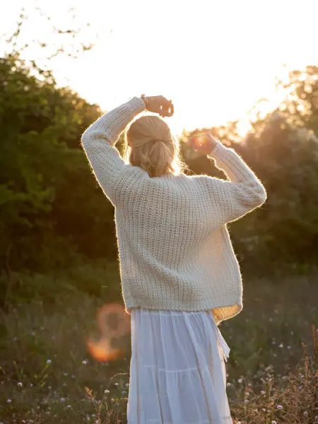 Dame med hvit kjole og hvit ulljakke står i et skogsområde med ryggen vendt mot kamera, og ser på solnedgangen.