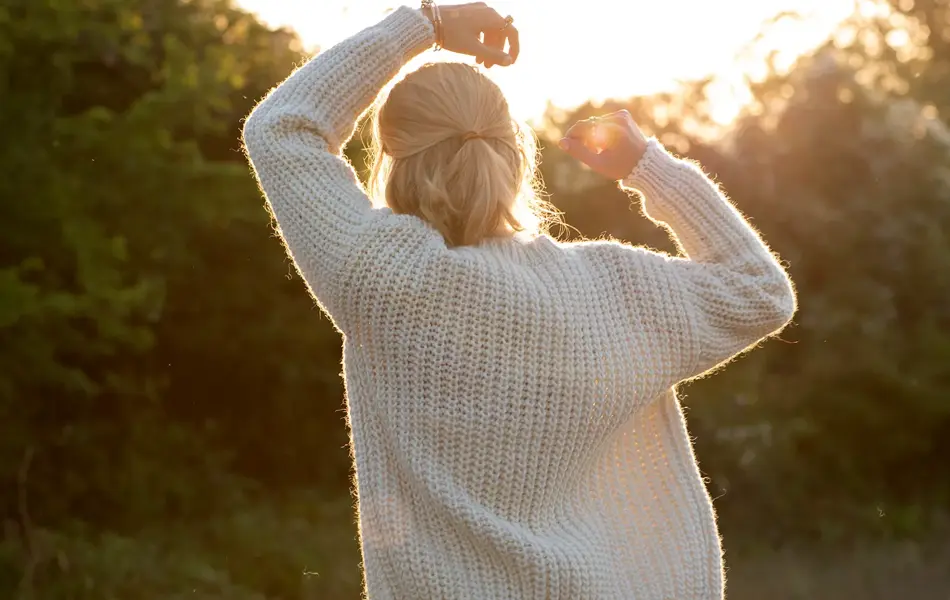 Dame med hvit kjole og hvit ulljakke står i et skogsområde med ryggen vendt mot kamera, og ser på solnedgangen.