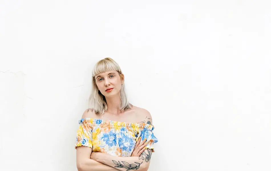 Dame med store bukser i denim og blomstermønstret topp står lent inntil hvit murvegg og ser mot kamera.