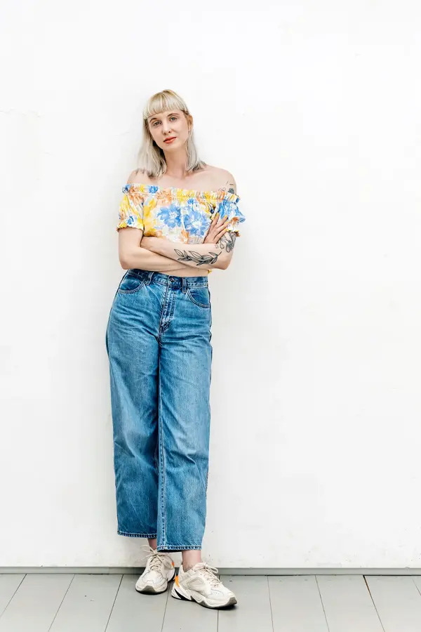 Dame med store bukser i denim og blomstermønstret topp står lent inntil hvit murvegg og ser mot kamera.