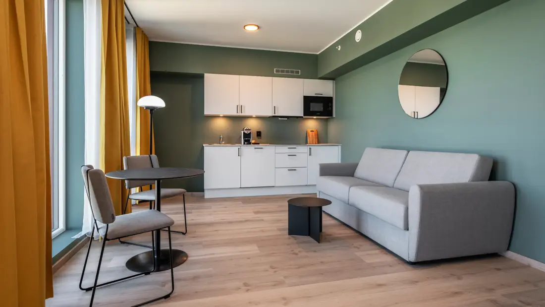 Business rom med kjøkken krok, grå sofa, spiseplass, grønne vegger, gule gardiner og parkett.