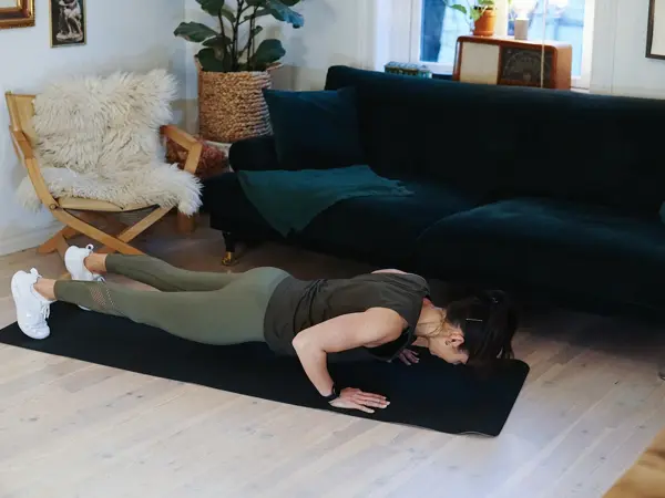 treningsekspert Anna Johansen trener på matte på gulvet, mørkegrønn sofa i bakgrunnen, lys stol med pels oppi, str grønn plante, bilder på veggen