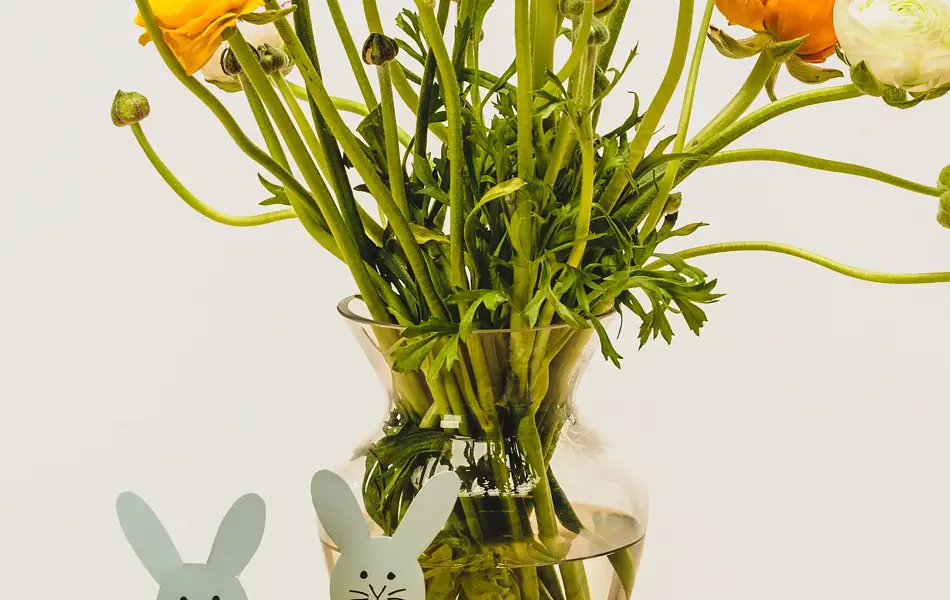 To grå kaniner med dusk laget av grønt og gult garn ved siden av en stor vase med blomster og sjokoladeegg på ett bord