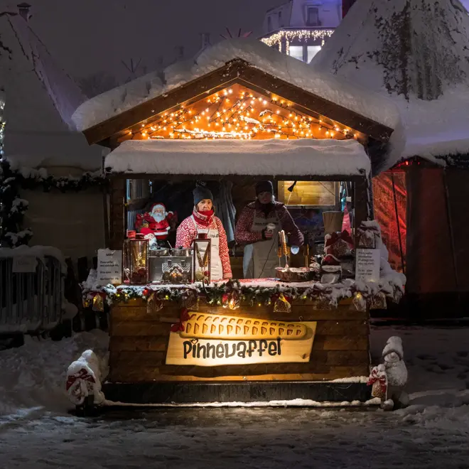 Julepyntet bod som selger pinnevaffel på Julemarkedet i Trondheim en snøfull vinterkveld. To selgere i strikkegensere står bak disken.