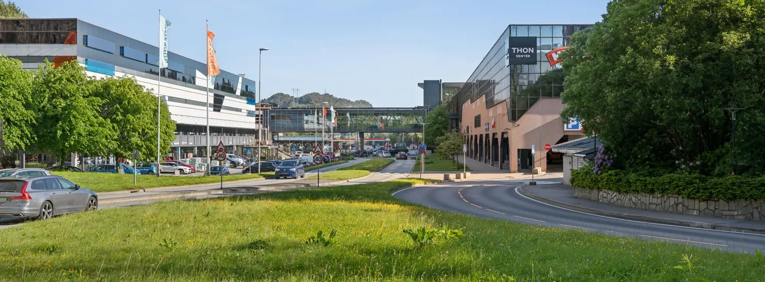 Eksteriørbilde av Thon Senter Vestkanten med kontor- og handelsbygg, gangbro mellom bygninger og grøntområde langs veien.