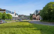 Eksteriørbilde av Thon Senter Vestkanten med kontor- og handelsbygg, gangbro mellom bygninger og grøntområde langs veien.