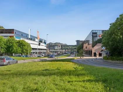 Eksteriørbilde av Thon Senter Vestkanten med kontor- og handelsbygg, gangbro mellom bygninger og grøntområde langs veien.
