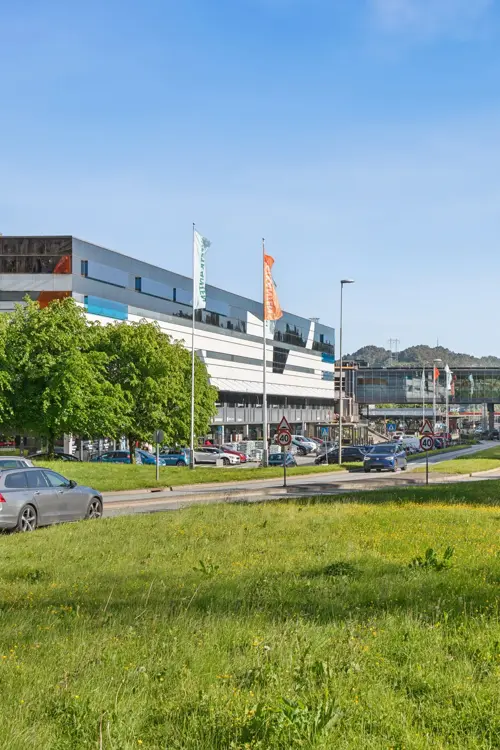Eksteriørbilde av Thon Senter Vestkanten med kontor- og handelsbygg, gangbro mellom bygninger og grøntområde langs veien.