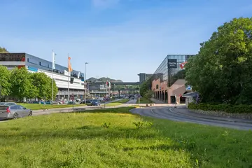 Eksteriørbilde av Thon Senter Vestkanten med kontor- og handelsbygg, gangbro mellom bygninger og grøntområde langs veien.