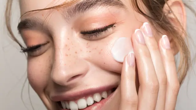Closeup studio shot of a beautiful young woman with freckles skin, applying moisturiser to her face. Posing against a grey background