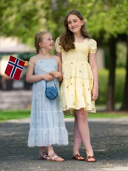 Liten jente i blå blomstrete lang kjole, liten blå veske og sandaler, holder et flagg, ved siden av større jente med gul kjole og brune sandaler.