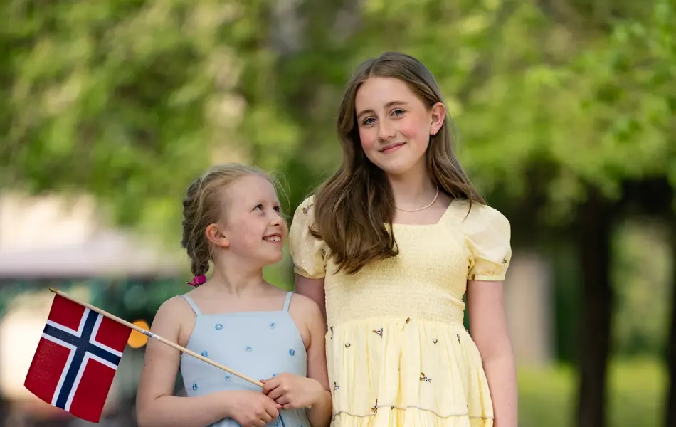 Liten jente i blå blomstrete lang kjole, liten blå veske og sandaler, holder et flagg, ved siden av større jente med gul kjole og brune sandaler.