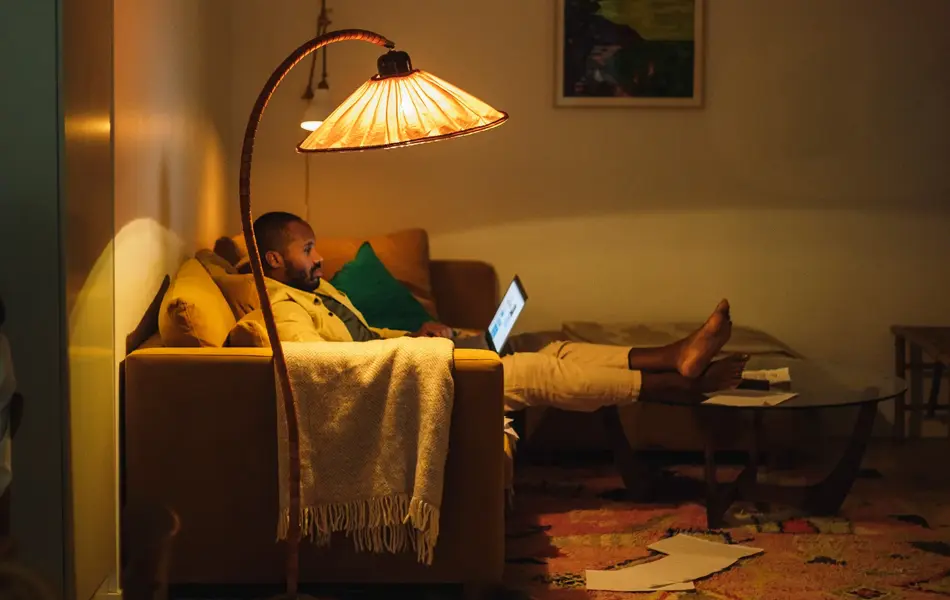 Mann sitter i sofa med pcen på fanget under lyset av en gulvlampe 