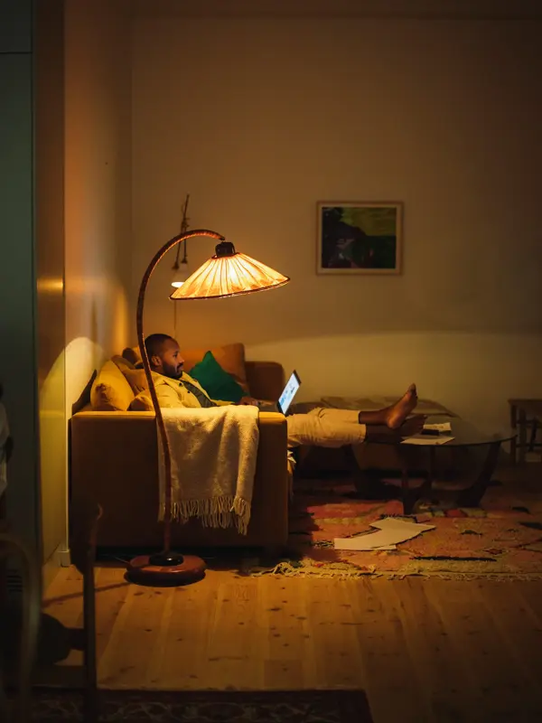 Mann sitter i sofa med pcen på fanget under lyset av en gulvlampe 