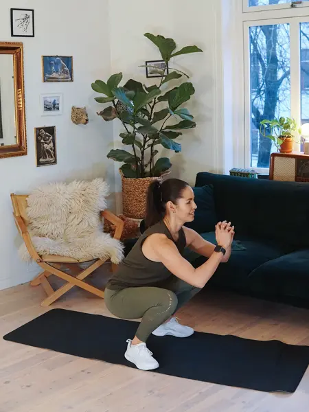 treningsekspert Anna Johansen trener på matte på gulvet, mørkegrønn sofa i bakgrunnen, lys stol med pels oppi, str grønn plante, bilder på veggen