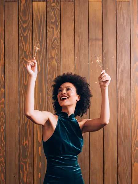 Smilende dame med blå fløyelskjole står foran en trevegg og holder opp to tente stjerneskudd