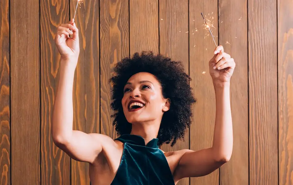 Smilende dame med blå fløyelskjole står foran en trevegg og holder opp to tente stjerneskudd