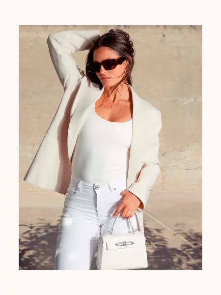 Dame står ute med hvit dressjakke, hvit singlet, hvite jeans og liten hvit veske.