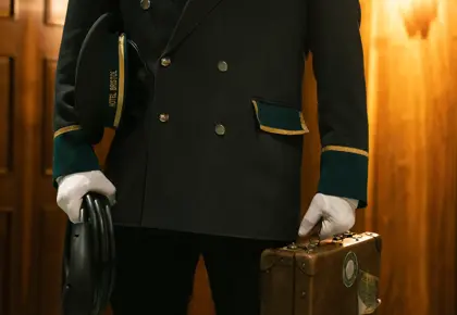 En ansatt i uniform og hvite hansker holder en koffert venstre hånd og en ladekabel til el-bil i høyre hånden.