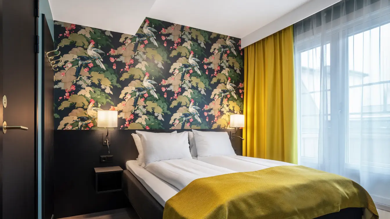 Blomstrete tapetvegg, vindu fra gulv til tak, gule gardiner, dobbeltseng, svart sengegavle med nattbord. Parkettgulv.