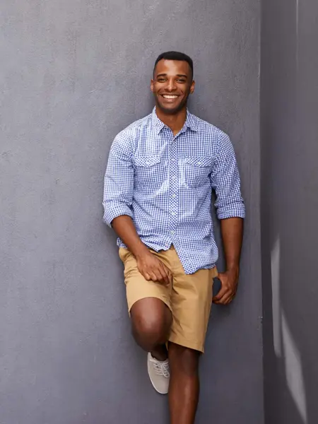 Mann i bermudashorts og skjorte som lener seg mot grå vegg